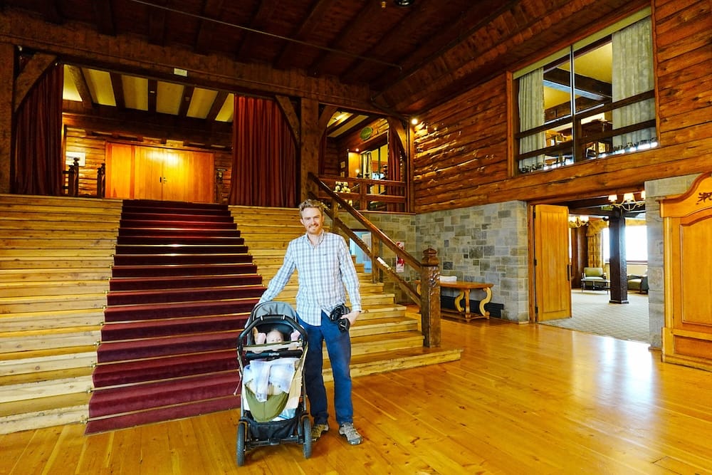 Samuel Jeffery de pie junto a su hija Aurelia en su cochecito dentro del lobby rústico del Hotel Llao Llao en Bariloche, Argentina. Se observa la arquitectura de troncos de ciprés, una gran escalera con alfombra roja y el estilo clásico de los refugios patagónicos.