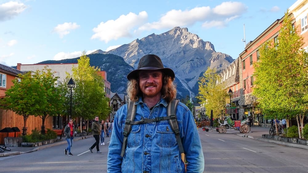 Samuel Jeffery parado en Banff Avenue en el centro de Banff, con peatones, vidrieras, un carruaje tirado por caballos y Cascade Mountain elevándose al final de la calle, Parque Nacional Banff, Alberta, Canadá.