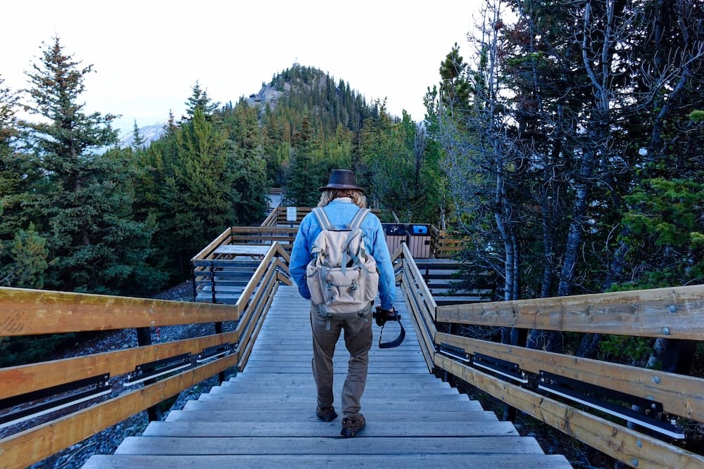 Samuel Jeffery on the Banff Gondola boardwalk toward Sanson's Peak, Sulphur Mountain, Banff, Alberta, Canada Samuel Jeffery walks away from the camera along the wooden boardwalk toward Sanson's Peak, backpack and camera in hand, surrounded by alpine forest on Sulphur Mountain near Banff, Alberta, Canada.