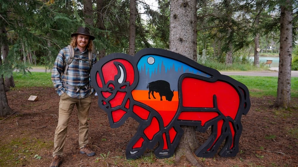 Bison sculpture installation on the Art in Nature Trail, Banff, Alberta, Canada Samuel Jeffery stands beside a large painted bison sculpture with a mountain and moon design along the Art in Nature Trail in Banff, Alberta, Canada.