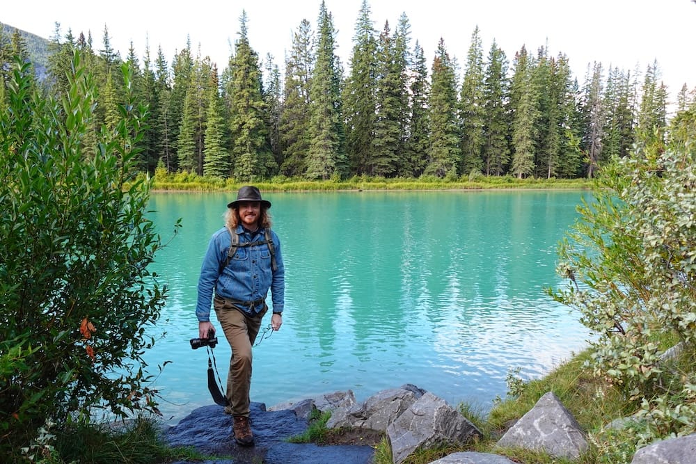 Samuel Jeffery stands on rocks beside a calm, turquoise stretch of the Bow River near the Banff Canoe Club, framed by forest near Banff, Alberta, Canada.