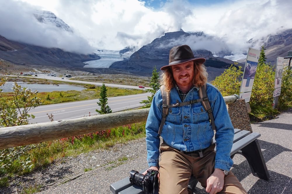 Samuel Jeffery sentado en un banco en un mirador del Columbia Icefield con una cámara, la ruta Icefields Parkway y el glaciar Athabasca visibles detrás de él, Alberta, Canadá.