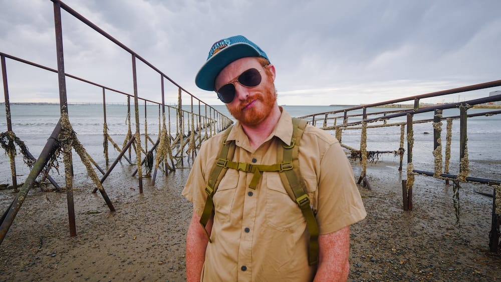 Samuel Jeffery standing among rusting sea defenses in Comodoro Rivadavia, Chubut, Argentina, highlighting the rugged Atlantic Patagonia coastline with barnacle-covered structures, tidal flats, and windswept coastal architecture.
