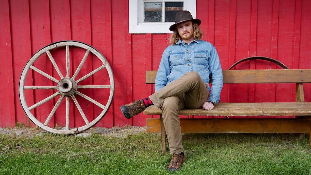 Samuel Jeffery at the covered wagon ride and cookout near Banff, Alberta Samuel Jeffery sits on a wooden bench beside an old wagon wheel leaning against a red barn wall during a covered wagon ride and cookout experience near Banff, Alberta, Canada.