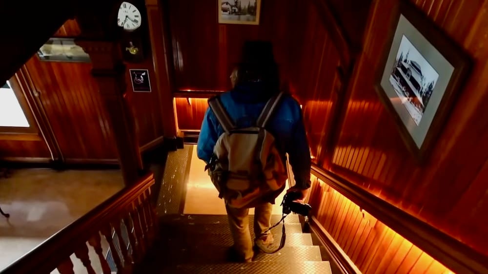 Samuel Jeffery, wearing a backpack and carrying a camera, walks down a wood-panelled staircase inside Banff Park Museum, past a vintage clock and framed historic photographs, in Banff, Alberta, Canada.