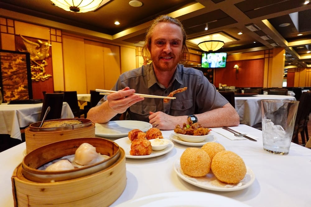 Samuel Jeffery with Dim Sum Spread at Silver Dragon Restaurant, Chinatown, Calgary, Alberta, Canada Samuel Jeffery holding chopsticks over a table of five dim sum dishes, including ginger beef and sesame balls, at Silver Dragon Restaurant in Chinatown, Calgary, Alberta, Canada.