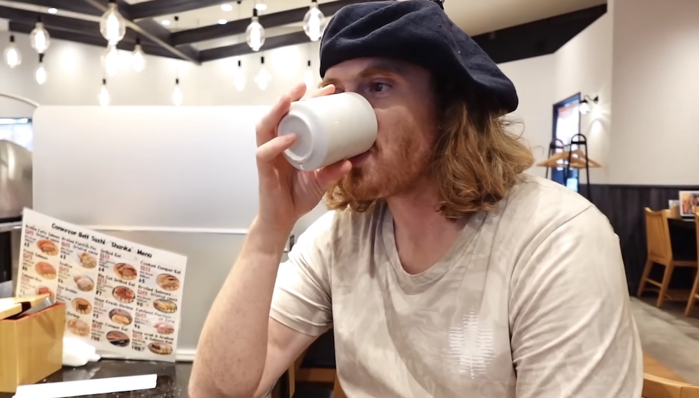 Samuel Jeffery Drinking Tea at a Casual Restaurant in Hakodate, Hokkaido, Japan, During a Sightseeing Break Samuel Jeffery drinks tea from a small white ceramic cup while sitting inside a casual restaurant in Hakodate, Hokkaido, Japan, wearing a black beret, with hanging lights and a food menu visible behind him.