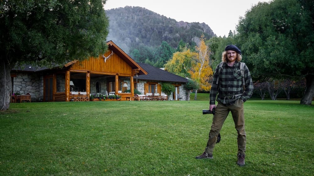 Samuel Jeffery stands on the lawn with a camera in front of the timber-and-stone main lodge at Estancia Arroyo Verde near Villa Traful, Neuquén, Argentine Patagonia, with autumn-colored trees and mountains behind.