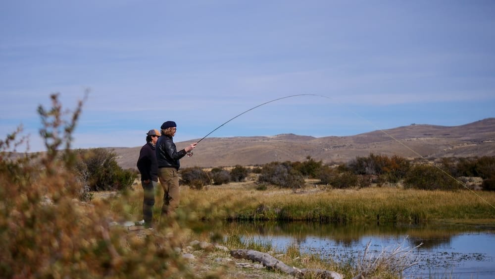 Samuel Jeffery fly fishes beside a small pond at Estancia Tecka in Chubut, Patagonia, Argentina, while a local guide watches his casting technique against open steppe grassland and low rolling hills.