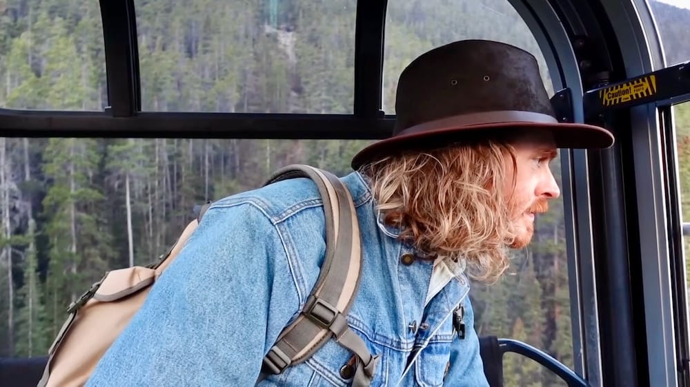 Samuel Jeffery looks out the window of a Banff Gondola cabin during the ride up, forested slopes visible outside, with no other visible seatmates in frame, Sulphur Mountain, Alberta, Canada.