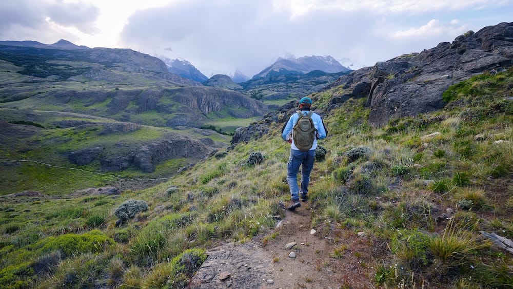 Samuel Jeffery caminando hacia el macizo del Fitz Roy cerca de El Chaltén, Santa Cruz, Argentina Samuel Jeffery caminando solo por un sendero de tierra a través de un valle rocoso y verde con una mochila pequeña, nubes de tormenta abriéndose para dejar ver el macizo nevado del Fitz Roy más adelante, en una caminata autoguiada cerca de El Chaltén, Santa Cruz, Patagonia argentina.