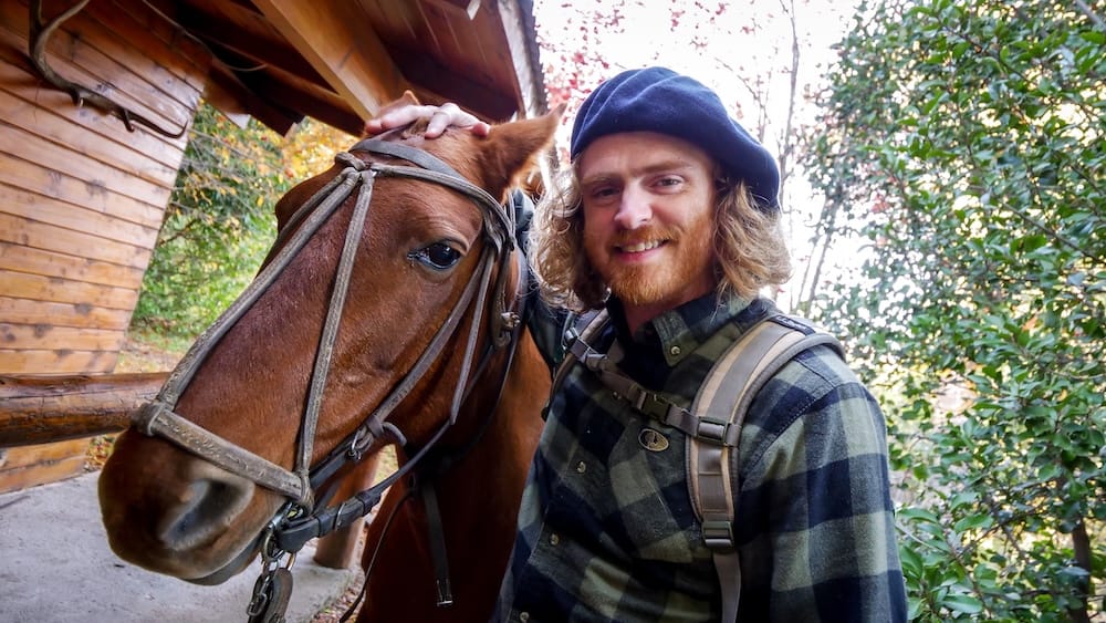 Samuel Jeffery con boina junto a un caballo en Estancia Arroyo Verde, Villa Traful, Neuquén, Argentina Samuel Jeffery con una boina azul marino y una camisa de leñador a cuadros, de pie junto a un caballo alazán con bridas tradicionales de cuero bajo el alero de madera de un galpón, follaje otoñal detrás, en Estancia Arroyo Verde cerca de Villa Traful, Provincia de Neuquén, Patagonia argentina.