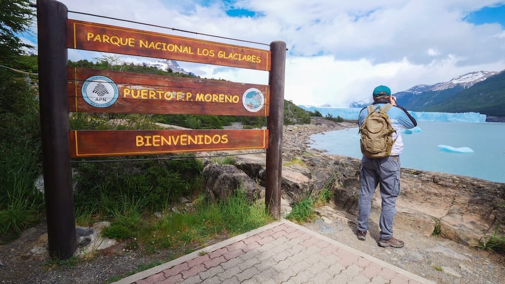Samuel Jeffery photographs Perito Moreno Glacier independently beside the Puerto F. Moreno sign in Los Glaciares National Park, Santa Cruz, Patagonia, Argentina, with turquoise water and floating ice behind him.