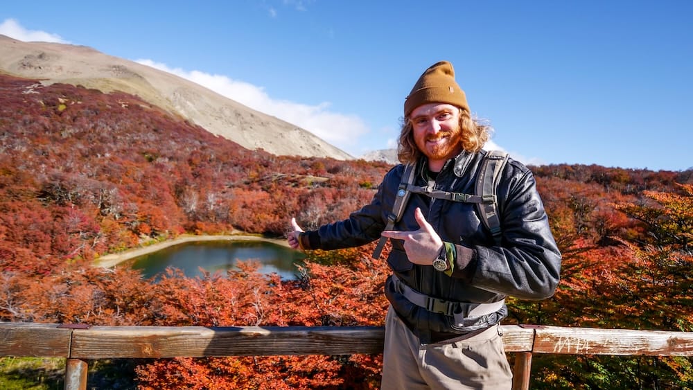 Samuel Jeffery overlooking Lago Gutiérrez during an autumn hike in Río Negro, Patagonia, Argentina Samuel Jeffery stands at a wooden viewpoint above Lago Gutiérrez, surrounded by vivid red and orange autumn foliage, a small mountain lake and pale rocky slopes in Río Negro, Patagonia, Argentina.