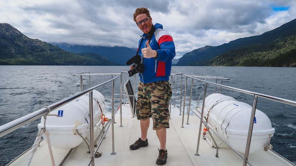 Samuel Jeffery standing on a boat in Los Alerces National Park, Argentina, with strong Patagonian winds visible in his hair while crossing glacial lake waters surrounded by Andean mountains and dense forest.