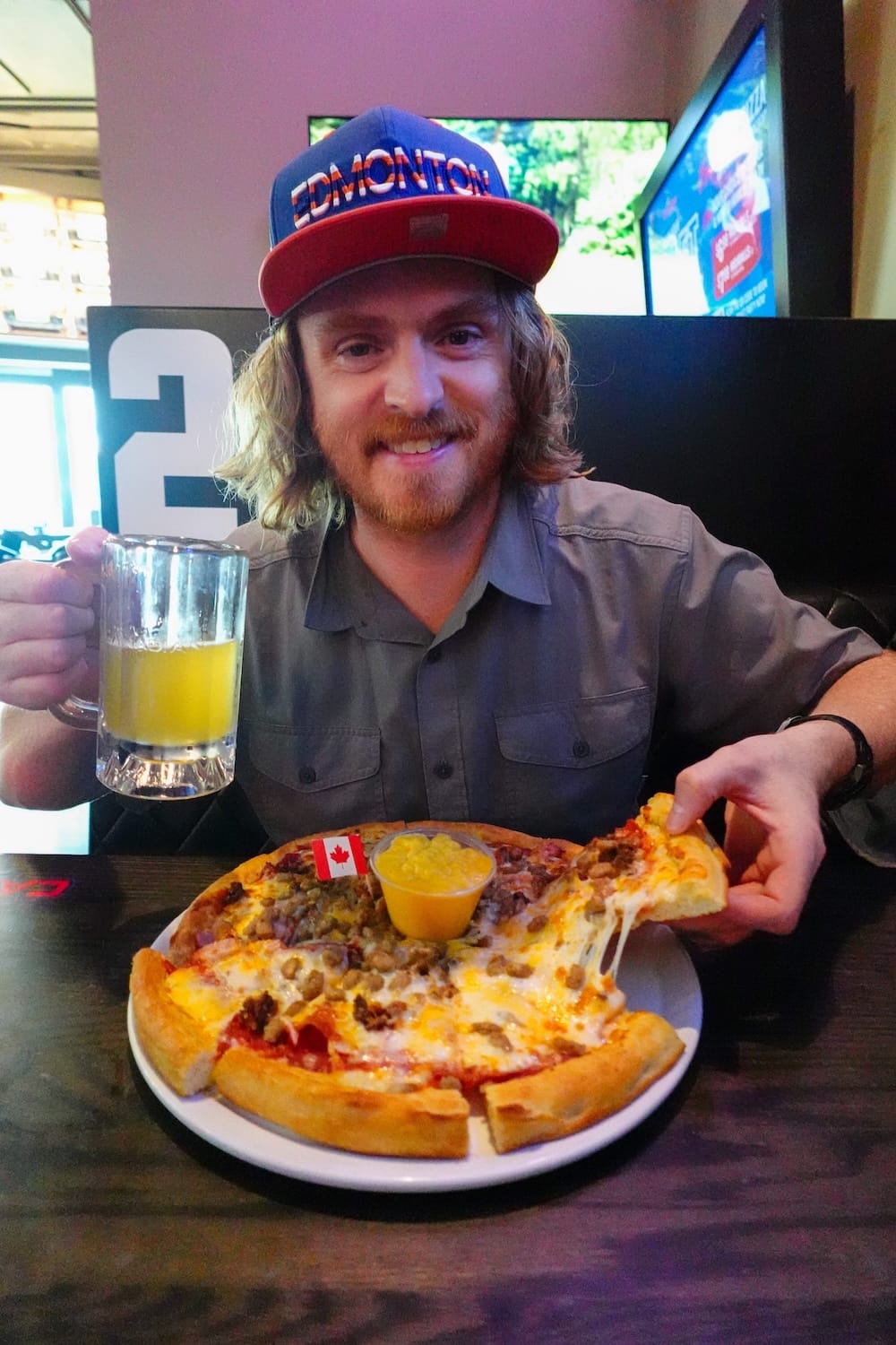 Samuel Jeffery with meaty pizza and beer at a sports bar, ICE District, Edmonton, Alberta, Canada Samuel Jeffery in an Edmonton cap holding a beer and lifting a slice from a meaty pizza with a side of nacho cheddar dip at a sports bar in the ICE District, Edmonton, Alberta, Canada.