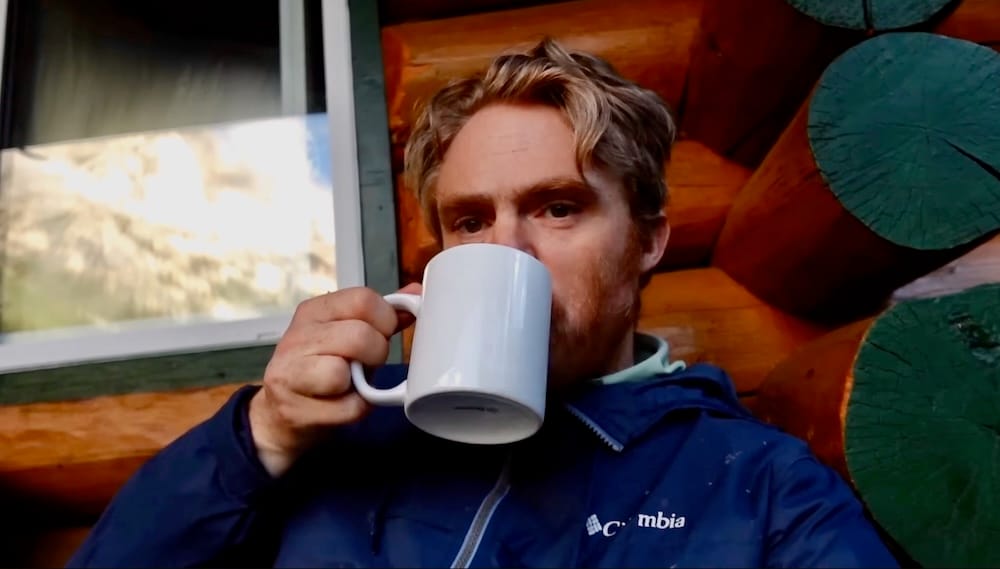 Samuel Jeffery, morning coffee, backcountry lodge, Banff National Park, Alberta, Canada Samuel Jeffery sips coffee outside a log backcountry lodge in Banff National Park, Alberta, Canada, with mountain scenery visible through a nearby window during a six-day horseback trek.