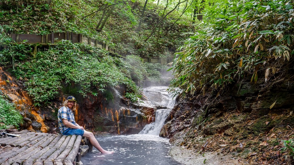 Samuel Jeffery en el baño natural de pies del río Oyunuma, Noboribetsu Onsen, Hokkaido, Japón, junto a una cascada humeante Samuel Jeffery sentado en una plataforma de madera con los pies sumergidos en el baño natural del río Oyunuma, en Noboribetsu Onsen, Hokkaido, Japón, junto a una pequeña cascada humeante y abundante vegetación del bosque.
