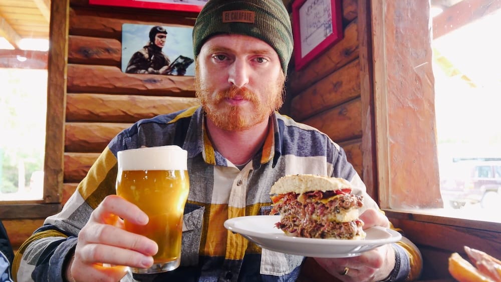 Samuel Jeffery sostiene una cerveza artesanal patagónica dorada y una gran hamburguesa gourmet dentro de un restaurante rústico de madera en El Calafate, Santa Cruz, Patagonia argentina.