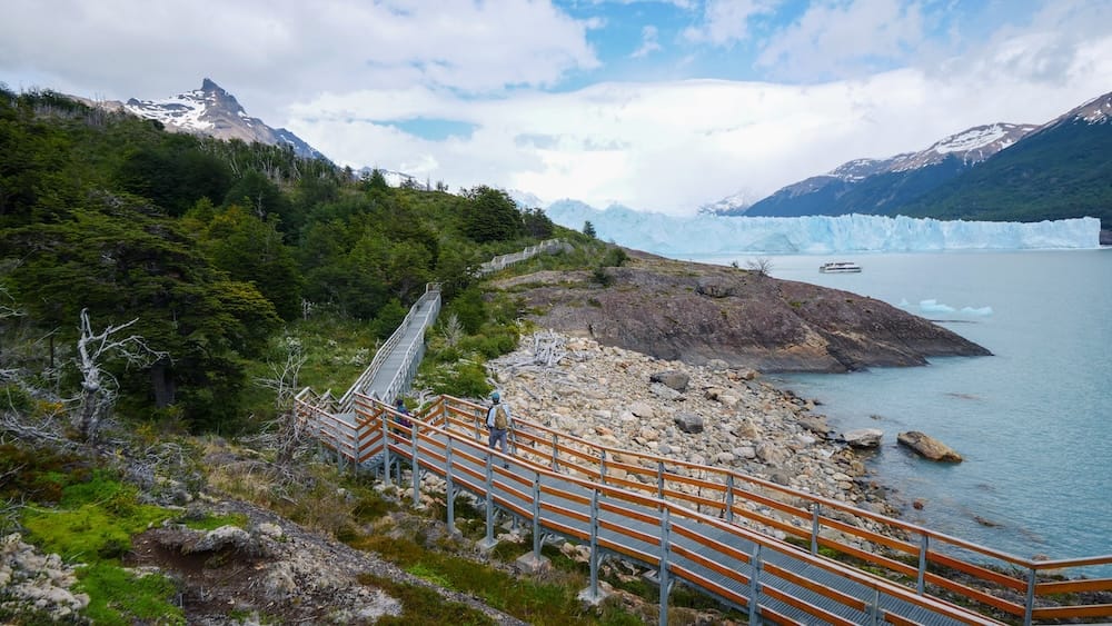 Samuel Jeffery on the Perito Moreno Glacier boardwalks in Santa Cruz, Patagonia, Argentina Samuel Jeffery walks along the elevated boardwalk stairs beside Perito Moreno Glacier in Santa Cruz, Patagonia, Argentina, with forest, rocky shoreline, lake water, a tour boat and the vast ice wall ahead.