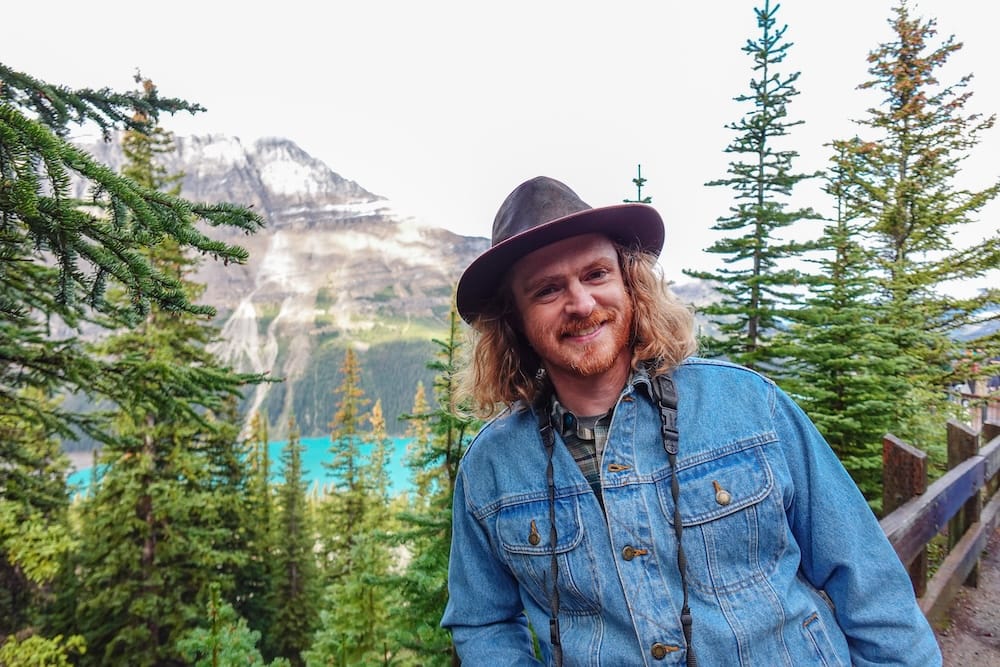 Samuel Jeffery at the Peyto Lake viewpoint, Icefields Parkway, Banff National Park Samuel Jeffery smiles beside evergreen trees at the Peyto Lake viewpoint along the Icefields Parkway in Banff National Park, Alberta, with the lake's turquoise water and a snow-streaked mountain visible behind him.