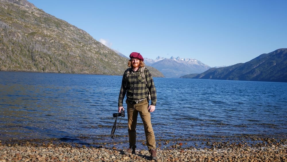 Photographer on the Shore of Lago Puelo, Chubut Province, Northern Patagonia, Argentina — Samuel Jeffery Samuel Jeffery stands on a pebbled shoreline holding a camera at Lago Puelo in Chubut Province, Northern Argentine Patagonia, with deep blue water, forested mountains, and snow-capped peaks framing the lake behind him.