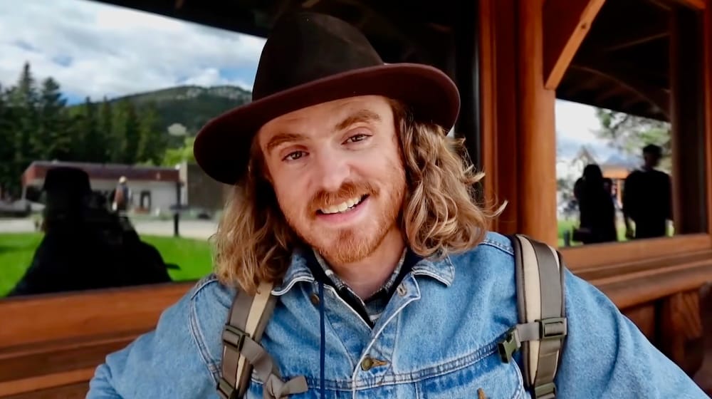 Samuel Jeffery smiling in a denim jacket and hat on the covered wood veranda outside Banff Park Museum, with mountains visible in the background, Banff, Alberta, Canada.