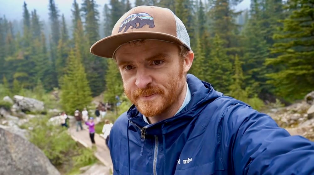 Samuel Jeffery takes a selfie on the Rockpile Trail at Moraine Lake in Banff National Park, Alberta, Canada, with a line of visitors visible on the stepped path behind him.