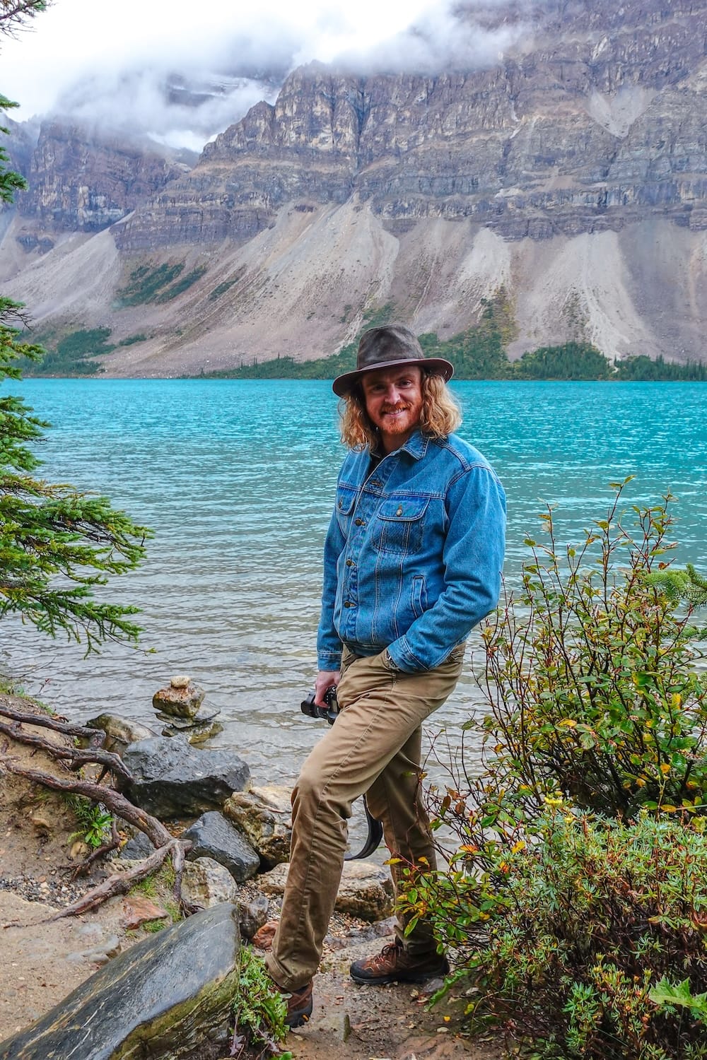 Samuel Jeffery poses on a rocky section of Bow Lake's shoreline along the Icefields Parkway in Banff National Park, Alberta, camera in hand, with vivid turquoise water and a steep, cloud-topped mountain behind him.