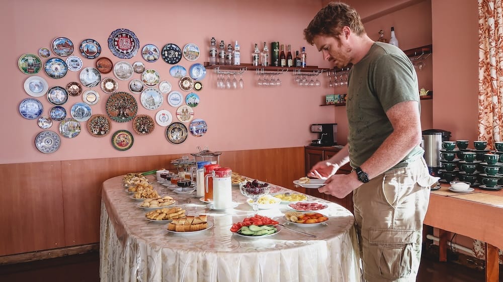 Samuel Jeffery selects food from a generous Kyrgyz breakfast spread featuring bread, pastries, sliced fruit, preserves, dairy drinks and tea, with decorative plates displayed on the guesthouse dining-room wall.
