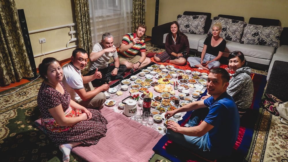 Samuel Jeffery sits with a local Kyrgyz family and friends around a large floor spread filled with tea, bread, salads, dumplings, fruit and shared dishes during a traditional home feast in Kyrgyzstan.

