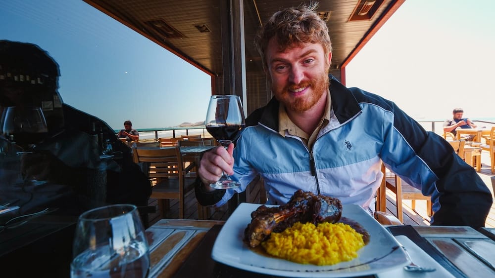 Slow-Roasted Lamb at a Seaside Restaurant, Rada Tilly, Chubut Province, Argentina — Samuel Jeffery Samuel Jeffery raises a glass of red wine at an open-air seaside restaurant in Rada Tilly, Chubut Province, Argentina, seated before a plate of slow-roasted Patagonian lamb and yellow rice with the beach and ocean visible behind him.
