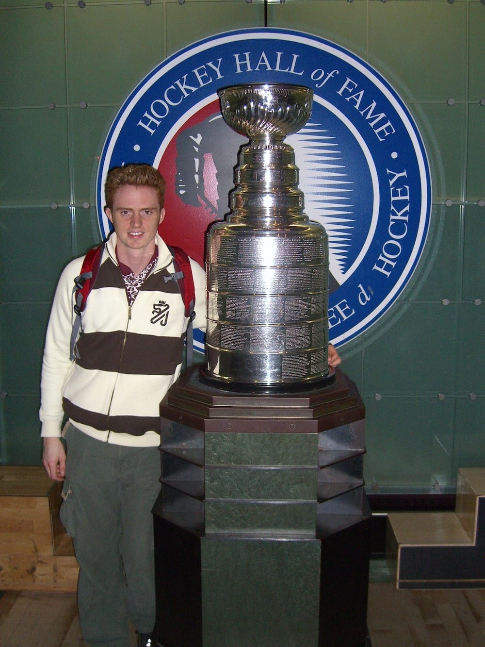 A younger Samuel Jeffery, sporting a 90s/2000s hairstyle, poses beside the Stanley Cup on display at the Hockey Hall of Fame in Toronto, Ontario, Canada, with the museum's circular logo visible on the wall behind him.