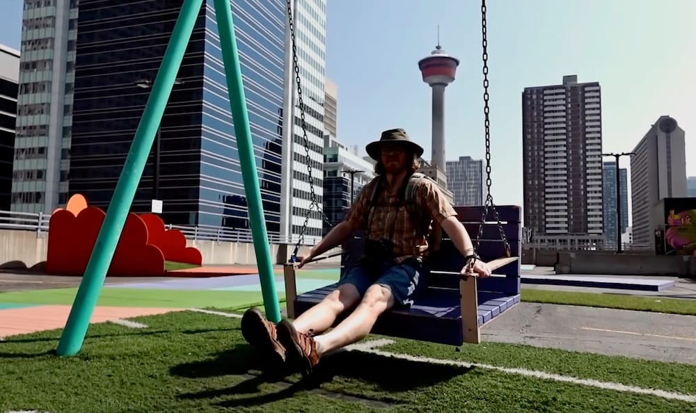 Samuel Jeffery sentado en una gran hamaca azul en la terraza de High Park en Calgary, Alberta, Canadá, con la Calgary Tower y el horizonte de la ciudad asomando atrás, y una escultura de una nube roja sobre el pasto sintético ahí cerca.