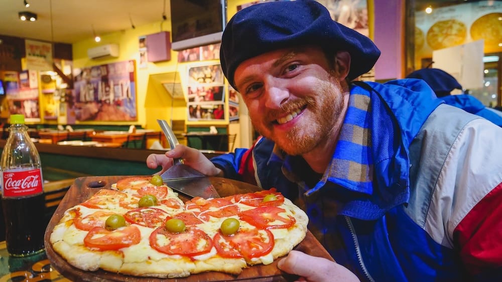 Samuel Jeffery eating tomato pizza in Esquel Patagonia inside cozy mountain pizzeria with Coca-Cola and thick crust Samuel Jeffery enjoying a tomato and cheese pizza in Esquel Chubut Argentina inside a cozy mountain pizzeria with olives and sliced tomatoes on a thick crust alongside a glass bottle of Coca-Cola in Patagonia.
