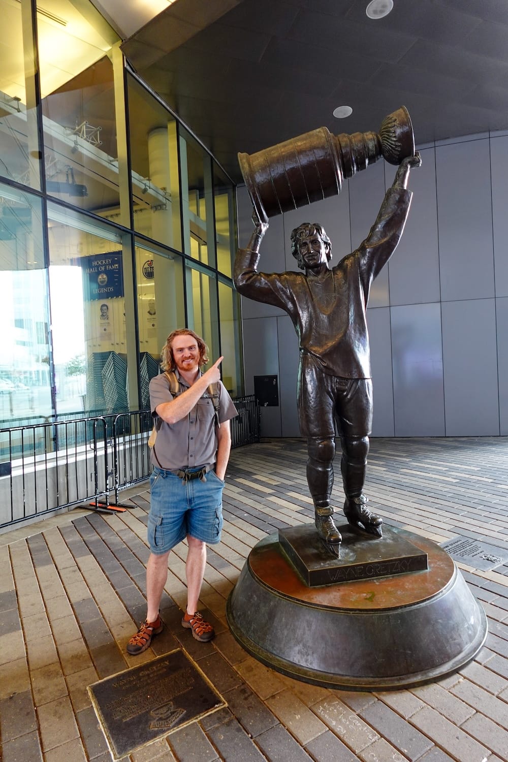 Samuel Jeffery points up at the bronze Wayne Gretzky statue holding the Stanley Cup overhead outside Rogers Place, with a Hockey Hall of Fame Legends banner visible in the background, Ice District, Edmonton, Alberta, Canada.