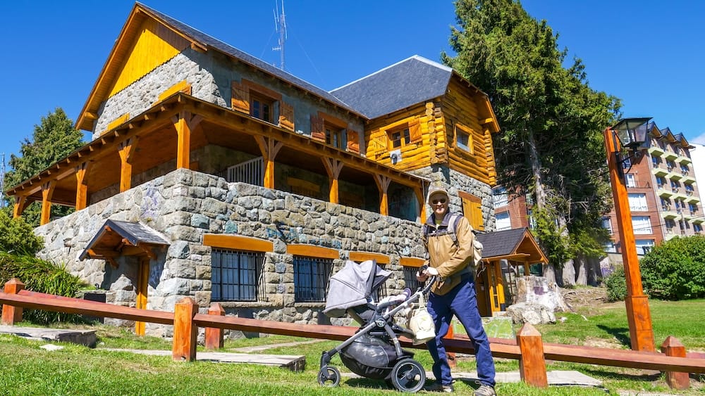 Samuel and Aurelia beside stone-and-wood architecture in Bariloche, Río Negro, Patagonia, Argentina Samuel Jeffery stands with Aurelia Jeffery in a stroller beside a rustic stone-and-log building in San Carlos de Bariloche, Río Negro, Patagonia, Argentina, beneath a clear blue sky.