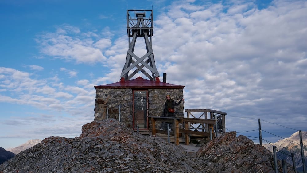 Audrey Bergner en el Observatorio Meteorológico de Sanson’s Peak, Sulphur Mountain, Banff, Alberta, Canadá Audrey Bergner junto al histórico Observatorio Meteorológico de Sanson’s Peak, en Sulphur Mountain sobre Banff, Alberta, Canadá, con terreno rocoso, barandas de madera y montañas bajo un cielo parcialmente nublado.