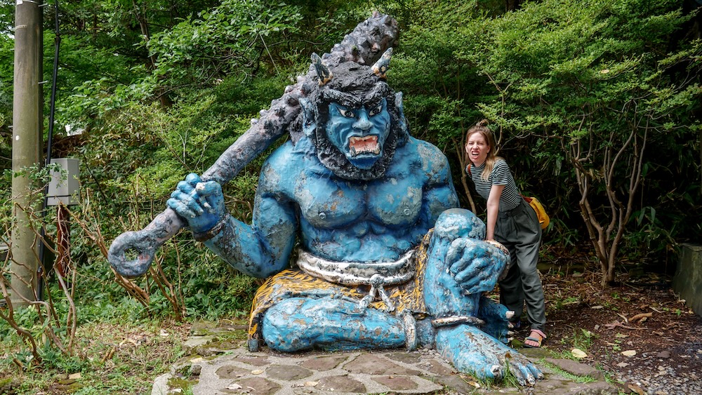 Audrey Bergner junto a un oni azul sentado con un garrote de púas en Noboribetsu Onsen, Hokkaido, Japón Audrey Bergner posando junto a una gran estatua de un oni azul sentado en Noboribetsu Onsen, Hokkaido, Japón. El demonio tiene cuernos, la boca abierta con colmillos afilados y sostiene un garrote con púas mientras está rodeado de árboles verdes.