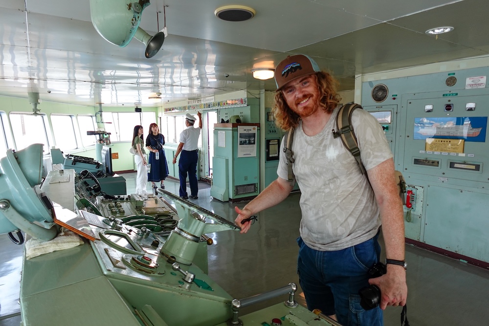 Samuel Jeffery Exploring the Bridge and Historic Navigation Controls of Seikan Ferry Memorial Ship Mashu-maru in Hakodate Samuel Jeffery smiling beside the pale green ship steering controls on the bridge of the Seikan Ferry Memorial Ship Mashu-maru in Hakodate, Hokkaido, Japan, with navigation equipment, large windows and other visitors behind him.