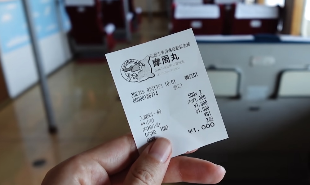 Admission ticket receipt aboard Mashu-maru, Seikan Ferry Memorial Ship, Hakodate, Hokkaido, Japan, with Audrey Bergner Audrey Bergner's hand holding a printed Japanese admission receipt for two adult tickets aboard the Seikan Ferry Memorial Ship Mashu-maru in Hakodate, Hokkaido, Japan, with blurred red passenger cabin seats in the background.