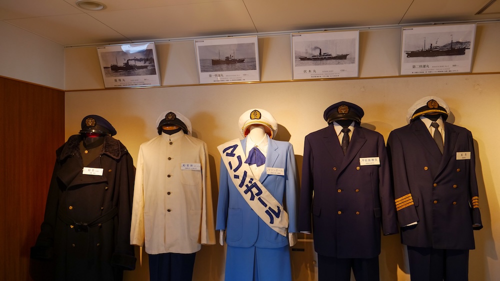 Crew uniform display and ferry photographs aboard Mashu-maru, Seikan Ferry Memorial Ship, Hakodate, Hokkaido, Japan Five preserved crew and captain uniforms with peaked caps and a blue Marine Girl suit with a white sash on mannequins below framed black-and-white ferry photos inside the Seikan Ferry Memorial Ship Mashu-maru in Hakodate, Hokkaido, Japan.