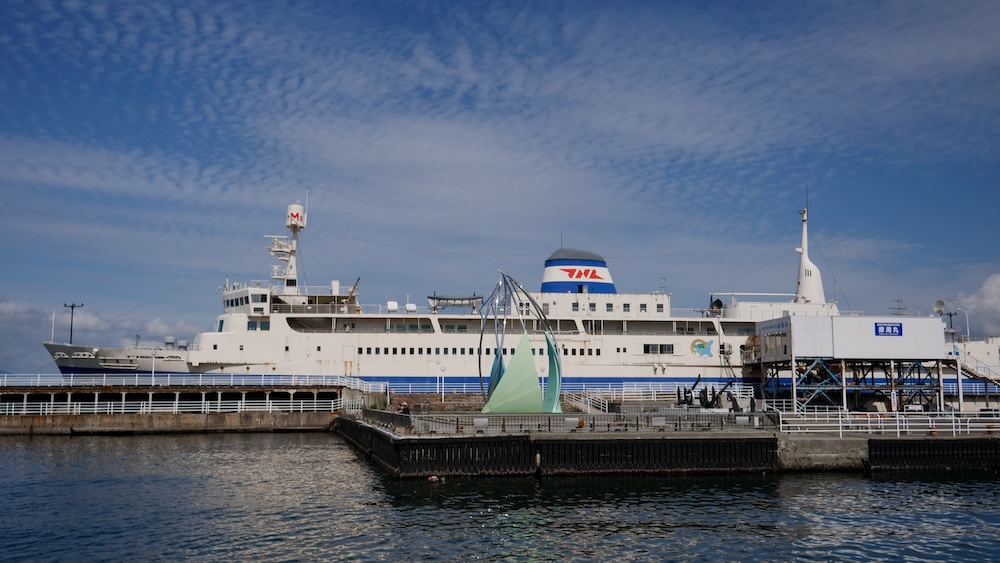Seikan Ferry Memorial Ship Mashu-maru moored at the harbor quay in Hakodate, Hokkaido, Japan White-and-blue hull of the Seikan Ferry Memorial Ship Mashu-maru moored at its harbor quay in Hakodate, Hokkaido, Japan, viewed in profile across the water with a curved green waterfront sculpture and elevated boarding walkway in front.