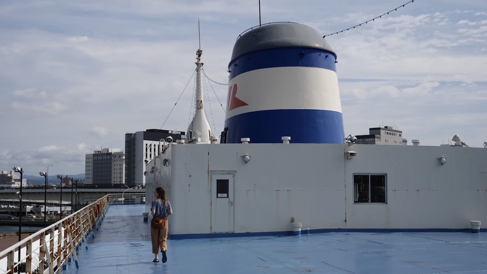 Audrey Bergner walking on the open deck of Mashu-maru, Seikan Ferry Memorial Ship, Hakodate, Hokkaido, Japan Audrey Bergner walking along the blue outdoor deck of the Seikan Ferry Memorial Ship Mashu-maru in Hakodate, Hokkaido, Japan, beside the white railing, blue-and-white smokestack funnel, and JR Inn and city buildings behind the harbor.