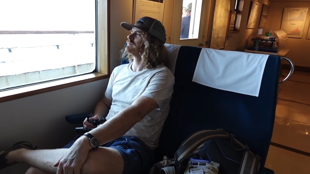 Preserved passenger seating aboard Mashu-maru, Seikan Ferry Memorial Ship, Hakodate, Hokkaido, Japan, with Samuel Jeffery Samuel Jeffery sitting in a dark-blue passenger seat with white headrest covers and looking out a cabin window on the Seikan Ferry Memorial Ship Mashu-maru in Hakodate, Hokkaido, Japan, with exhibition panels and a display case behind him.