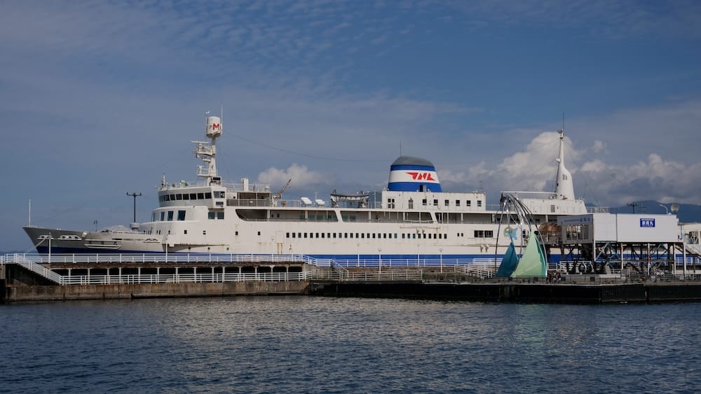 Seikan Ferry Memorial Ship Mashu-maru docked on the Hakodate waterfront in Hokkaido, Japan, with the preserved white railway ferry, terminal structures and harbour water visible under a blue sky.