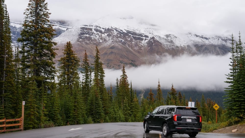 Una SUV oscura estacionada a un lado de la ruta mojada en la Icefields Parkway cerca de Peyto Lake, con nieve fresca cubriendo las montañas arriba y niebla baja pasando por el valle, Alberta, Canadá.