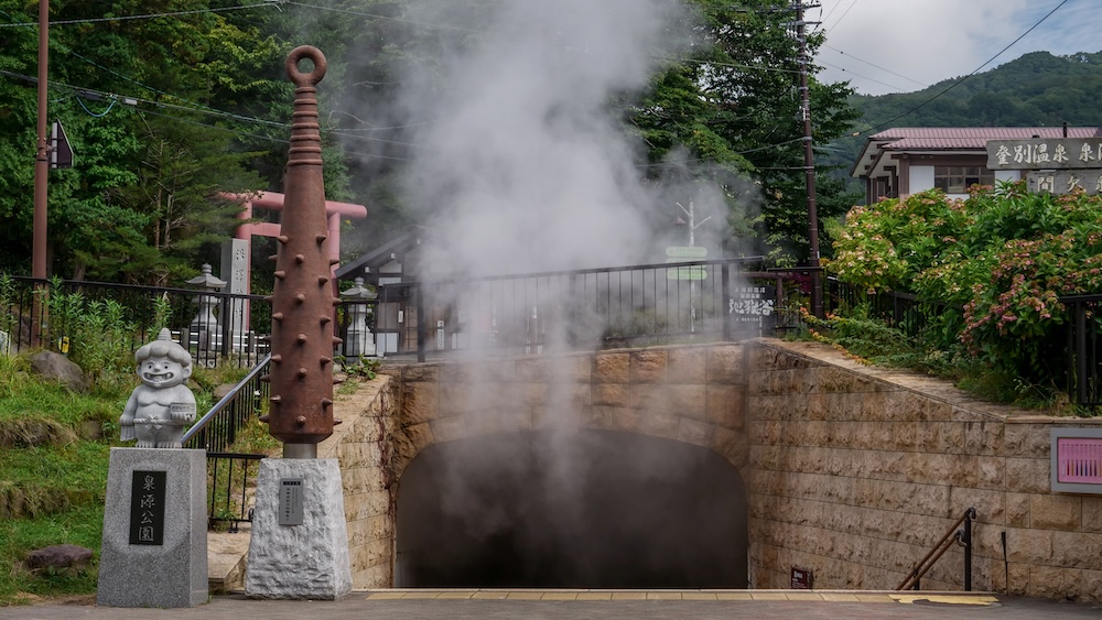 Géiser geotérmico del parque Sengen expulsando vapor junto a una estatua oni en Noboribetsu Onsen, Hokkaido, Japón Una densa columna de vapor blanco sale del géiser del parque Sengen, dentro de un recinto de piedra en Noboribetsu Onsen, Hokkaido, Japón. A su lado hay un monumento alto con púas y una pequeña estatua oni, con árboles y edificios detrás.