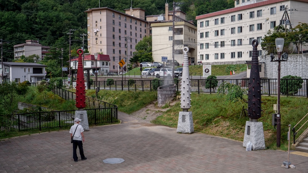 Géiser del parque Sengen y esculturas gigantes de garrotes de oni en Noboribetsu Onsen, Hokkaido, Japón Garrotes de oni gigantes en rojo, blanco y negro sobre pedestales de piedra alrededor del géiser del parque Sengen, con una columna de vapor y hoteles termales en la ladera detrás, en Noboribetsu Onsen, Hokkaido, Japón.