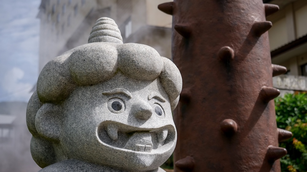 Primer plano de la estatua gris de piedra tallada del demonio Onikko, con un solo cuerno y una sonrisa con colmillos, junto a un alto garrote kanabo marrón con púas y el vapor del géiser en el parque Sengen de Noboribetsu Onsen, Hokkaido, Japón.